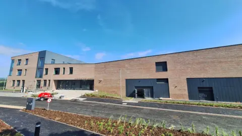 A wide, modern school building set under a bright blue sky with light, wispy clouds.