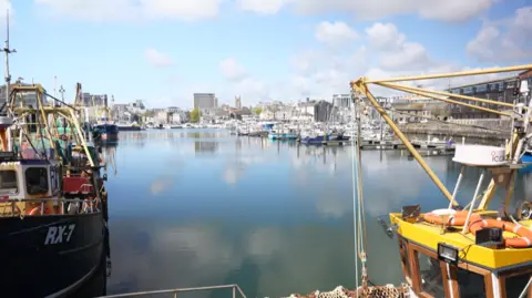Boats in a quay in Plymouth. The sky is blue with clouds and there is a reflection of the sky in the water. There are tall buildings in the background some distance away.