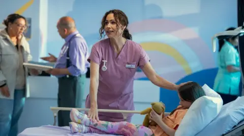 Run Away Ingrid Greene, played by Minnie Driver, is wearing pink hospital scrubs as she stands over a young girl, who is clutching a teddy bear, in a hospital bed on a ward. 