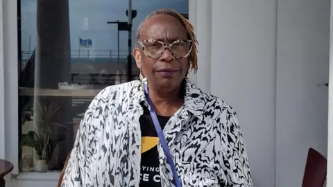 Fiona McCarthy A black woman in a black and white jacket and glasses, looking towards the camera. A white wall with a large window is seen in the background.