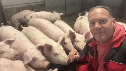 Alistair Lane Farmer Alastair Lane, with short dark hair, wearing a red hooded jumper and a red and black boiler suit, kneeling down with a number of pigs surrounding him