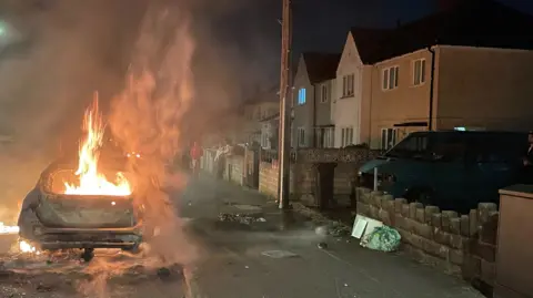 A car which is on fire. It is parked next to a pavement with a row of house on the rigth hand side.