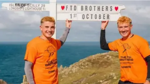 Laura Newbury Photography Two men in orange t-shirts standing in front of the sign for Land's End, on a cliff top overlooking the sea. The t-shirts are printed with a logo of a running brain, with The FTD Brothers written underneath. The Land's End sign has been customised to say "FTD Brothers, 1st October".