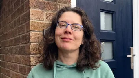 Anna stood outside her front door. It is a close crop image. She is smiling, with brown curly hair down to her shoulders, wearing glasses and a light green hoodie.