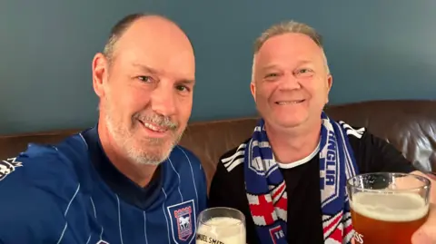 Supplied Two men in a selfie-style image smiling at the camera. They are holding pints of beer in their hands and wearing matching blue football shirts.