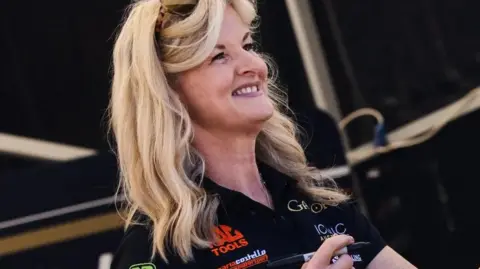 IOM TT Maria Costello, who has long blond hair an is wearing a branded T-shirt, smiling while signing autographs.
