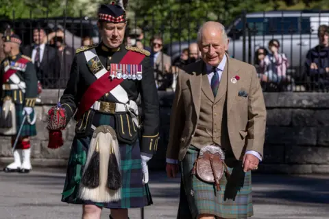 PA Media King Charles III walks alongside a soldier in ceremonial Highland dress during a parade inspection. The King is wearing a tweed jacket, waistcoat and tartan kilt, while the soldier wears a dark dress uniform decorated with medals, a tartan kilt and traditional military accessories. Spectators watch from behind railings in the background. 