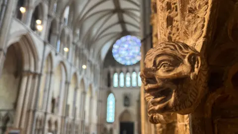 On one side of the image is a focused view of a carving of a 3D face inside of stone, to it's left is a blurred view of the inside of Lincoln Cathedral. Light seeps through a stained glass window with blue, pink and red hues in the background.