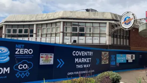 A large blue sign on a grey building which reads Coventry Market in yellow lettering