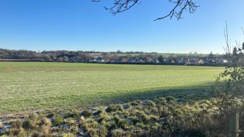 Amy Holmes/BBC A picture of a green field in Redbourn. There is a small amount of frost on the ground at the front of the picture and then a green field which has a row of houses behind it. The sky is blue behind the houses.