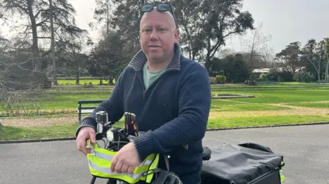 Ross Le Brun is looking at the camera, he is straddling his bike and the handlebars and part of the cargo section are visible. His feet are on the ground. The bike has neon yellow on the front to make it more visible. He is wearing a blue quarter-zip jumper with a green T-shirt underneath and has sunglasses on his head. Behind him part of a road, trees and a mowed lawn are visible.