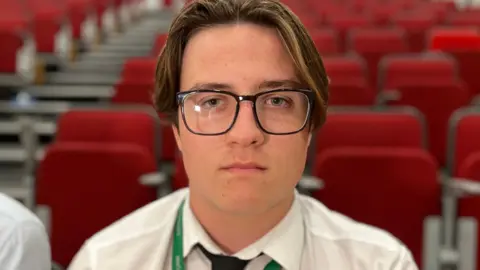 Charlie Rose/BBC In a white shirt, dark tie and dark-framed glasses, student James, who wears his hair in a centre parting, looks at the camera with rows of red empty theatre seats behind him.