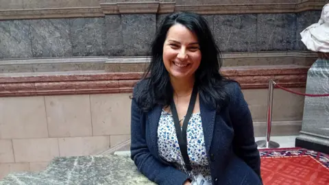 A woman with shoulder-length black hair is smiling and wearing a blue floral top and navy jacket. She is standing on a marble staircase in Sheffield Town Hall.