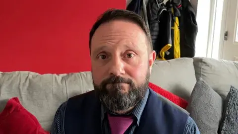 John Newcombe looks directly at the camera, he has dark brown hair and a beard and moustache, he is wearing a navy waistcoat with a blue pinstriped shirt and a pink tie. He is sitting on a grey sofa, the wall behind him is red.