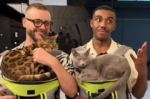 TfL A screenshot from the video, showing two men with two sleeping cats sat on cat carriers. The men are looking away from the camera, in mid-conversation.