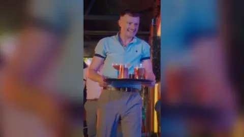 Man with short brown hair, wearing a blue shirt and grey trousers, carrying a tray of drinks.