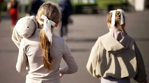 Two girls seen from behind wearing beige and cream clothing with white ribbons in their hair. One is holding a white rabbit soft toy