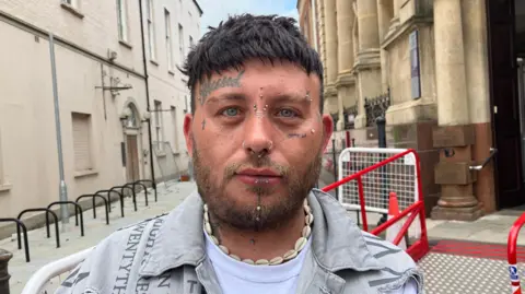 Headshot image of a man with facial tattoos and facial piercings, wearing a denim jacket and a shell necklace standing on a street