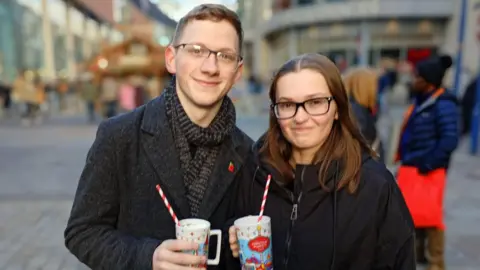 Leo Harvey and Amy Hulton, both wearing black coats, smile as they held up hot chocolates in Manchester Christmas markets mugs