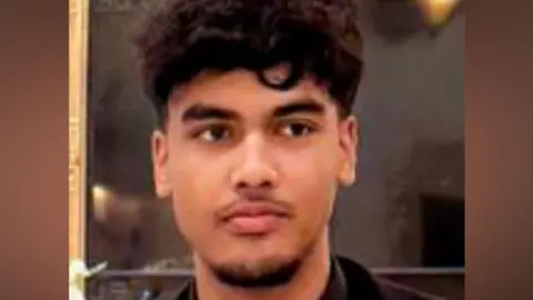 Absi Ahmed looking to the right of the camera lens. He has dark, curly hair which is short on the sides and long on the top. The photo is cropped close, with only his head and shoulders visible. He is wearing a black suit jacket, black shirt and black tie. He has a thin moustache and a beard covering just his lower chin.