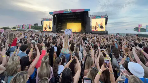 Music fans, some sitting on shoulders with hand-written signs, most filming ton their iphones, crown around the stage in Herrington Country Park. Zara Larsson is seen on large yellow screens wearing a blue dress and being held aloft by her dancers. 