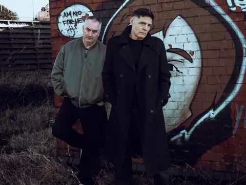 Soma Records Two men standing in front of a brick wall that has graffiti on it. One is wearing a black coat and black trousers while the other wears a khaki jacket and black trousers.