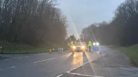 BBC A police car with its headlights on, blocking off the road. Two police officers can be seen wearing high vis yellow jackets. Yellow cones and a police warning sign are on the road in front of the car. 
