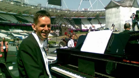 Iain Farrington Iain Farrington in black tie sitting at a piano in a stadium. There are people and camera crews in the background as well as vehicles with trailers.
