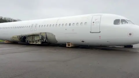 A large white plane fuselage which has had it's wings removed, leaving a gaping hole in the side of the plan. It is sitting on the tarmac runway at Cotswold Airport.