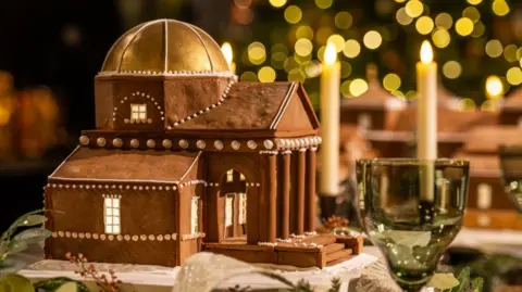 Emily Garland A gingerbread replica of a 18th Century Palladian-style country house. The gingerbread building has been topped off with icing and white edible beading to carve out the windows and other detailing. There is a dome on top of the building and there are also fairy lights in the background of the building. There are also two candles in the frame.