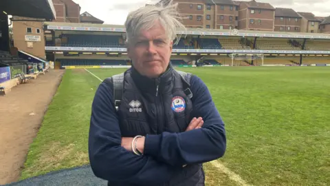 Warren Muggleton/BBC A man with his arms folded standing on the edge of a football pitch and a braintree town badge on his coat