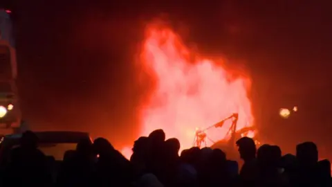 Red flames and silhouettes of people gathered behind the flames