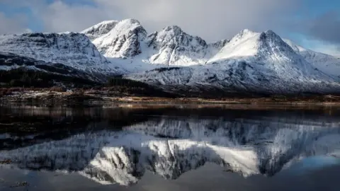 A mountain range covered with snow with the peaks reflected in a loch in the foreground