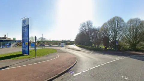 The entrance to a petrol station with trees to the left of the road