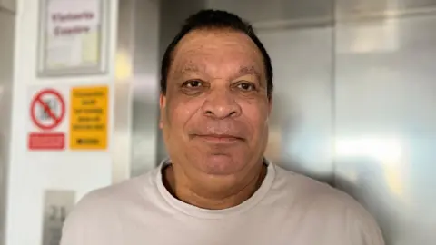 Oscar Smith, a 71-year-old mixed-race man with short black hair wearing a beige t-shirt and standing in front of a closed lift. 