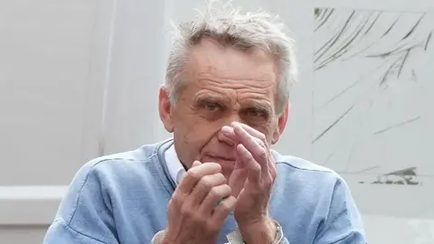 Eddie Mitchell An elderly man with his hands in front of his face in handcuffs.
