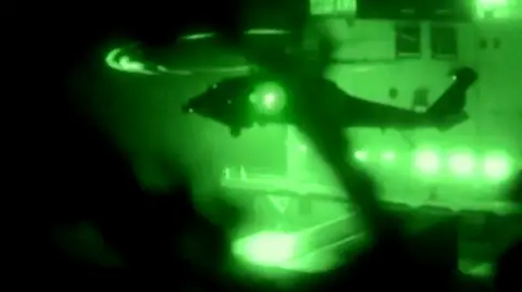 A helicopter hovers in front of a ship in footage shot at night