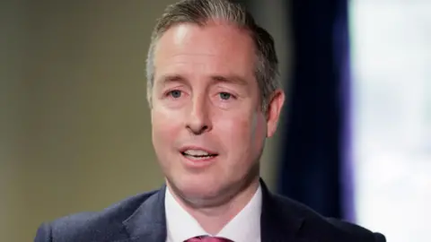 Shows head and shoulders of a man in a dark suit, white shirt and pink tie