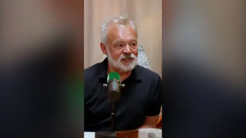 Graham Norton wearing a black polo shirt. He's sat in front of a green mic.