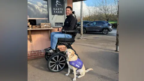 Man in a powered wheelchair. It lifts off the ground and is adjustable in height. There's a Labrador sat on the ground at a cafe outside.