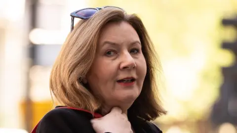 PA Media Teacher Rebecca Richardson has shoulder-length blonde/brown hair. She is wearing a black top and has her hand on a red bag strap and has sunglasses on her head. The background behind her is blurred.