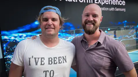 Two men stand opposite each other, embracing each other and looking into the camera.