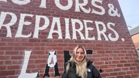 Blonde-haired 21 year old woman, in a black puffa jacket holding a paintbrush covered in white paint, in front of a brick terraced house in Sheffield where she is repainting a sign advertising a former slating company 