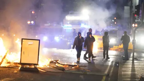 People wearing hoodies around their faces walk in the street next to burning debris in the middle of the road. There are police vehicles behind them.