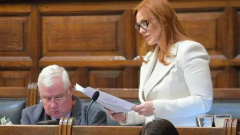 Claire Christian, a woman with ginger hair, she wears glasses and a white blazer as she stands up and reads a speech. 