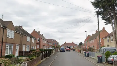 A quiet residential street lined with semi-detached brick houses on both sides. The homes have small front gardens with low fences, and some feature bay windows. Several cars are parked along the road and in driveways. Tall trees are visible on the right side, and overhead utility wires stretch across the cloudy sky.