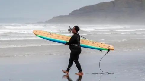 Deanna Bowen Jess Blake holding a foam board on her hip, she has the leash attached to her ankle and is wearing a wetsuit. In the background is the sea with a lot of white water waves and a headland jutting out into the sea. 