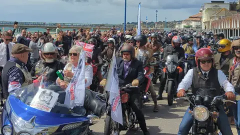 A huge group of motorcyclists are gathered by the sea in Bournemouth. Many of the bikers are smartly dressed and wearing ties. 