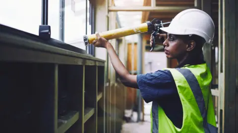 A woman is inside a building and is wearing a white helmet, blue T-shirt and yellow and silver fluorescent jacket. She is holding a caulk gun which has a yellow body and a long white nozzle. 