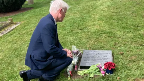 Paul James is kneeling by the plaque and placing flowers by it.